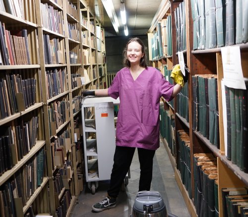 Person mit Staubsauger und Staubtuch im Magazin zwischen Bücherregalen